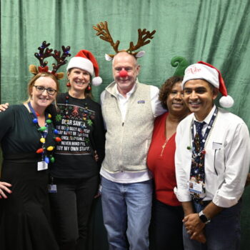 Five staff members posing and smiling together: two with reindeer antlers, two with Santa hats, and one sporting a green head prop. The central figure wears a red ball nose and antler prop.